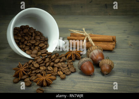 Roasted coffee beans spilling from a cup, acorns and cinnamon with star anise spice on a rustic wooden table in a close up view Stock Photo