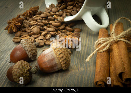 Roasted coffee beans spilling from a cup, acorns and cinnamon with star anise spice on a rustic wooden table in a close up view Stock Photo