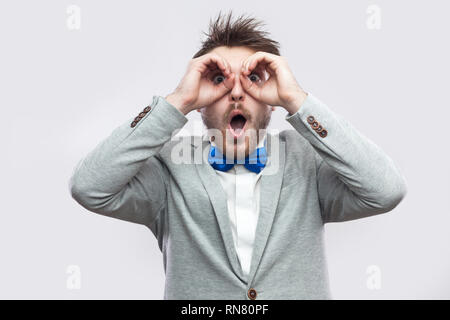 Portrait of surprised handsome bearded man in casual grey suit, blue bow tie standing with binoculars gesture hand on eyes and looking at camera. indo Stock Photo
