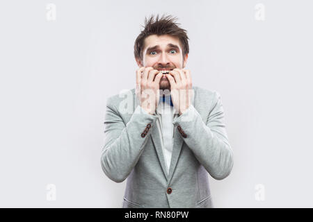 Portrait of worry handsome bearded man in casual grey suit and blue bow tie standing, nervous bitting his nails and looking at camera. indoor studio s Stock Photo