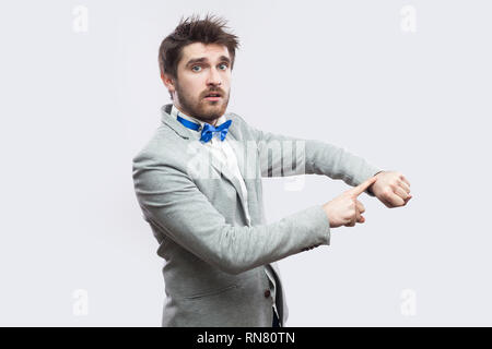 Portrait of serious worried handsome bearded man in casual grey suit and blue bow tie standing with watch hand gesture and looking at camera. indoor s Stock Photo
