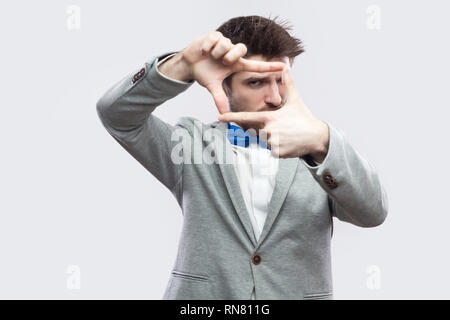 Portrait of serious handsome bearded man in casual grey suit and blue bow tie standing and looking at camera with cropping composition gesture. indoor Stock Photo