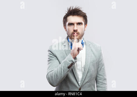 Portrait of serious handsome bearded man in casual grey suit and blue bow tie standing, looking at camera with silence sign. indoor studio shot, isola Stock Photo