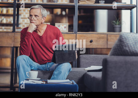 Thinking. Calm aged man holding a modern tablet and thoughtfully looking into the distance Stock Photo