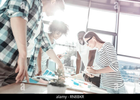 Team work. Cheerful partners standing around table and bowing heads while examining their project Stock Photo