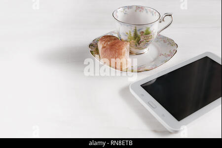 On a white surface we have a small plate and a porcelain cup, a half croissant and an electronic tablet. Copy space at the left side and hi key. Stock Photo