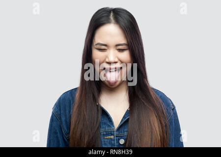 Portrait of funny crazy beautiful brunette asian young woman in casual blue denim jacket with makeup standing with closed eyes and tongue out. indoor  Stock Photo
