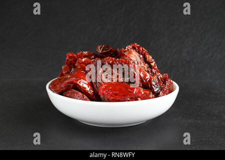 Sun-dried tomatoes in sunflower oil with spices (rosemary, garlic, basil, black pepper) in a white ceramic bowl, standing on a black stone surface aga Stock Photo