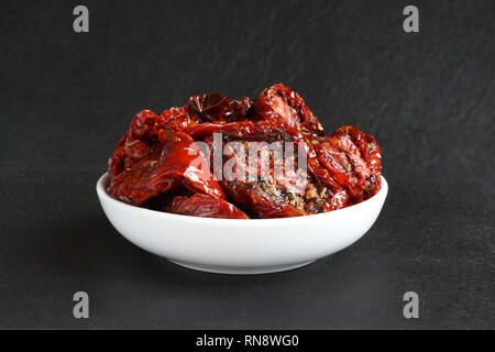 Sun-dried tomatoes in sunflower oil with spices (rosemary, garlic, basil, black pepper) in a white ceramic bowl, standing on a black stone surface aga Stock Photo