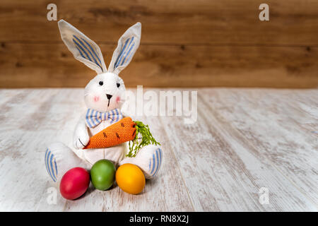 Close-up of three colorful Easter eggs in front of Easter bunny holding carrot on white painted and vintage oak background, front view Stock Photo