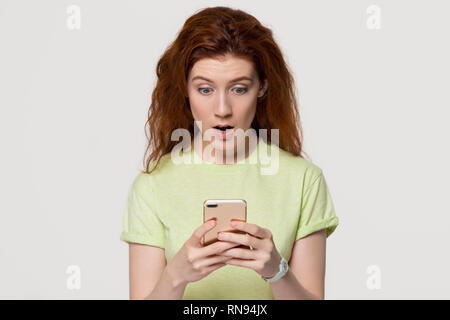 Shocked amazed redhead woman holding phone astonished about unexpected sms Stock Photo