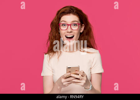 Excited redhead woman holding cell phone amazed by mobile win Stock Photo