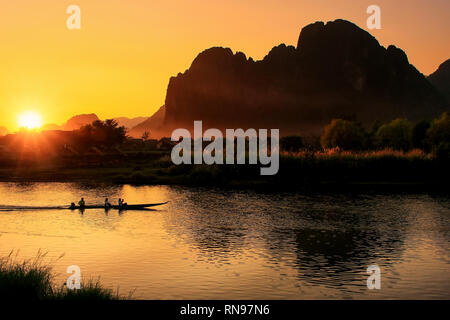 Sunset over Nam Song River with silhouetted rock formations and a boat in Vang Vieng, Laos. Vang Vieng is a popular destination for adventure tourism  Stock Photo