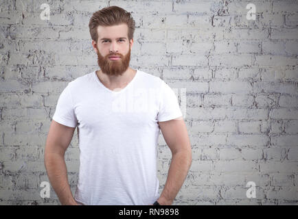 Serious thoughtful male with ginger beard, dressed casually, looking to the camera, isolated over white brick wall background. Stock Photo