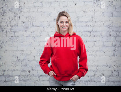 Pretty smiling young woman standing by the brick wall with her hand in the pockets in red sweatshirt. Stock Photo