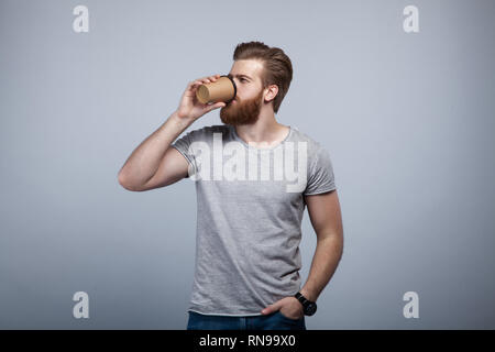 young handsome bearded man in grey t-shirt drinking his cup of coffee holding his hand in the pocket. Stock Photo