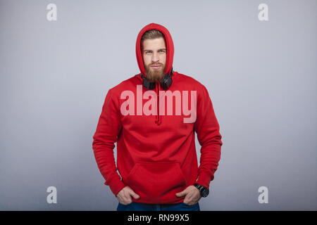 young handsome man with the beard wearing red sweatshirt with hood on and headphones on his neck standing at the grey background looking to the camera Stock Photo