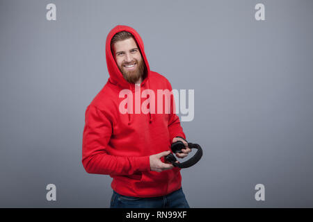 young handsome smiling bearded man in red hoodie on holding headphones in his hands standing at the grey background and looking to the camera. Stock Photo