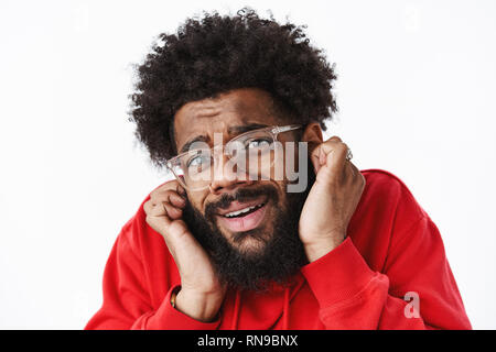 Headshot of bothered and upset african american timid guy with beard in glasses closing ears with index fingers frowning suffering headache as hearing Stock Photo