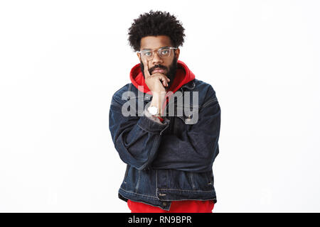 Serious-looking throughtful good-looking african-american hip-hop dancer with beard in glasses and denim jacket over hoodie touchign face with finger Stock Photo