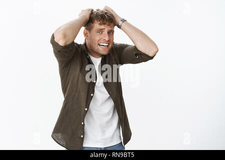 Intense distressed fair-haired handsome man watching horror feeling bad for person, holding hands on head, stooping grimacing from empathy and worry Stock Photo