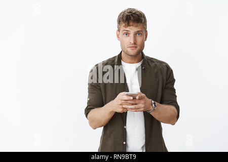 Waist-up shot of guy being distracted with question turning attention at camera while holding smartphone, using mobile phone, wasting time playing Stock Photo