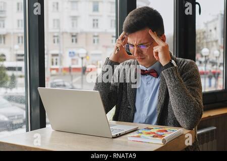 Young tired, ill, overworked man in formal wear sitting in front of computer and touching his forehead Stock Photo