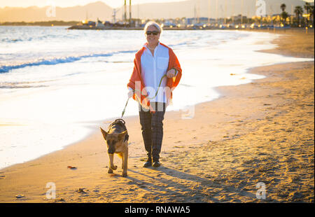 Beautiful retired older woman and pet german shepard dog walking along the shore sea ocean on beach in Companionship Benefits of animals Keeping activ Stock Photo