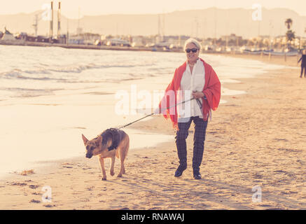 Beautiful retired older woman and pet german shepard dog walking along the shore sea ocean on beach in Companionship Benefits of animals Keeping activ Stock Photo