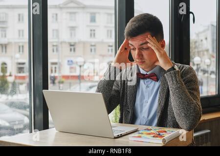 Young tired, ill, overworked man in formal wear sitting in front of computer and touching his forehead Stock Photo