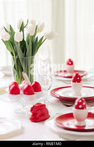 Happy easter. Decor and table setting of the Easter table is a vase with white tulips and dishes of red and white color. Easter colored eggs with whit Stock Photo