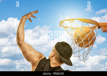 Man wearing black t-shirt and black cap throwing basketball ball into hoop on blue sky white clouds background Stock Photo
