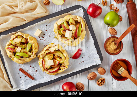 uncooked galettes with apple slices stuffing topped with butter cubes, prepared to bake on a baking tray. ingredients, rolling pin and cloth at the ba Stock Photo