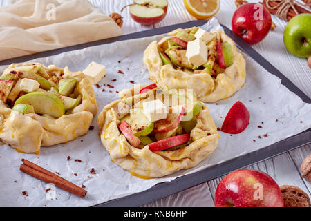 open apple pies, galettes with apple slices stuffing topped with butter cubes, prepared to bake on a baking tray. apple, cinnamon sticks and kitchen c Stock Photo