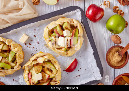 open apple pies, galettes with apple slices stuffing topped with butter cubes, prepared to bake on a baking tray. apple, cinnamon sticks and kitchen c Stock Photo