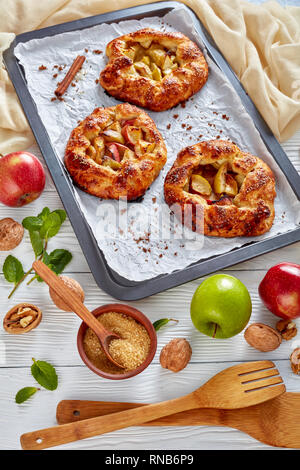 delicious open apple pies, galettes with apple slices stuffing on a baking tray. apple, cinnamon sticks, brown sugar and ivory kitchen cloth at the ba Stock Photo