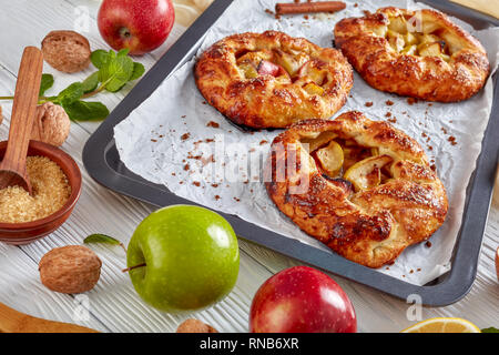 delicious open apple pies, galettes with apple slices stuffing on a baking tray. apple, cinnamon sticks, brown sugar and ivory kitchen cloth at the ba Stock Photo