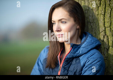 Unhappy Looking Young Woman Leaning Against Tree On Winter Walk In Park Stock Photo