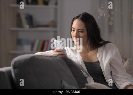 Serious woman checking smartphone content sitting on a couch in the night at home Stock Photo