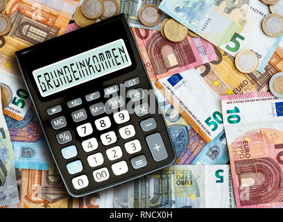German word GRUNDEINKOMMEN (basic income) written on display of pocket calculator against cash money on table Stock Photo