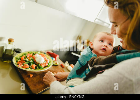 Smiling baby ported in baby carrier backpack looking at his mother while she cooks, concept of family conciliation Stock Photo