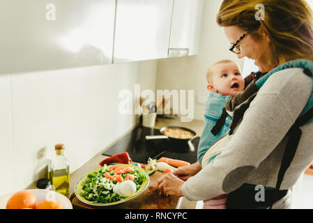 Smiling baby ported in baby carrier backpack looking at his mother while she cooks, concept of family conciliation Stock Photo