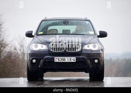 GRODNO, BELARUS - OCTOBER 2016: BMW X5 with glowing headlights standing on asphalt under rain covered with raindrops over gray rainy sky and forest ba Stock Photo