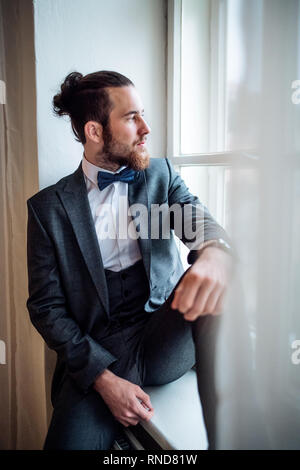 A handsome hipster young man with formal suit sitting on a window sill on an indoor party. Stock Photo