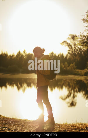 Young man walking beside a lake wearing a backpack on a sunny day. Traveler walking in a forest alone with sun in the background. Stock Photo