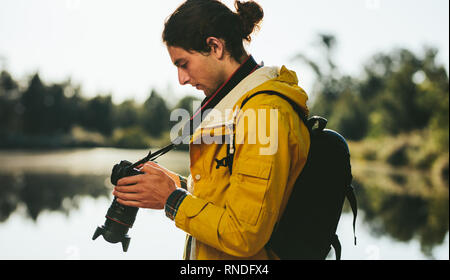 Side view of a tourist standing near a lake looking at his dslr camera. Close up of a traveler looking at his digital camera standing outdoors. Stock Photo