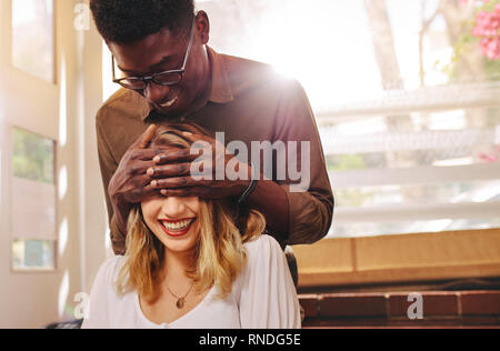 Young man standing behind his girlfriend and covering her eyes in cafe. Couple on date, man surprising woman at coffee shop. Stock Photo