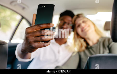 Couple sitting on back seat of a car and taking selfie. Focus on mobile phone in hand of man taking selfie with girlfriend in the car. Stock Photo