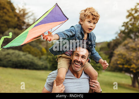 Little boy holding a kite sitting on his father's shoulders at the park. Happy father and son flying kite outdoors. Stock Photo