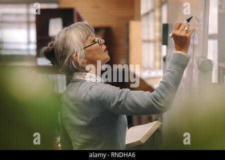 Side view of a senior female teacher writing on a white board in classroom. Close up of a woman lecturer teaching in classroom. Stock Photo